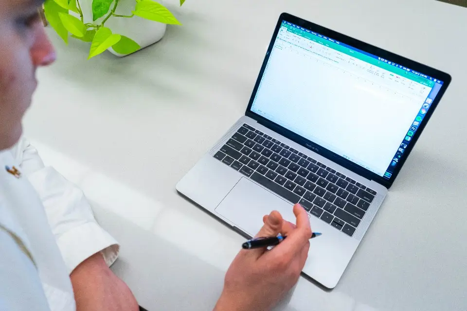Pessoa com caneta na mão diante de um notebook aberto sobre mesa branca