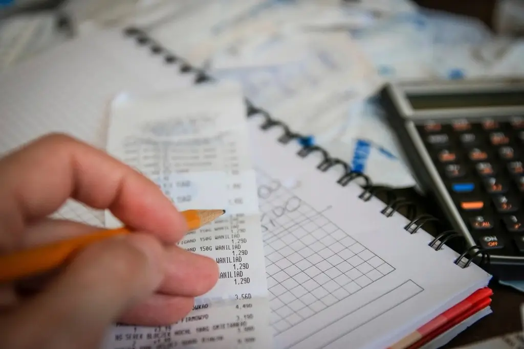 Mão com lápis conferindo recibos e anotações, com uma calculadora ao lado