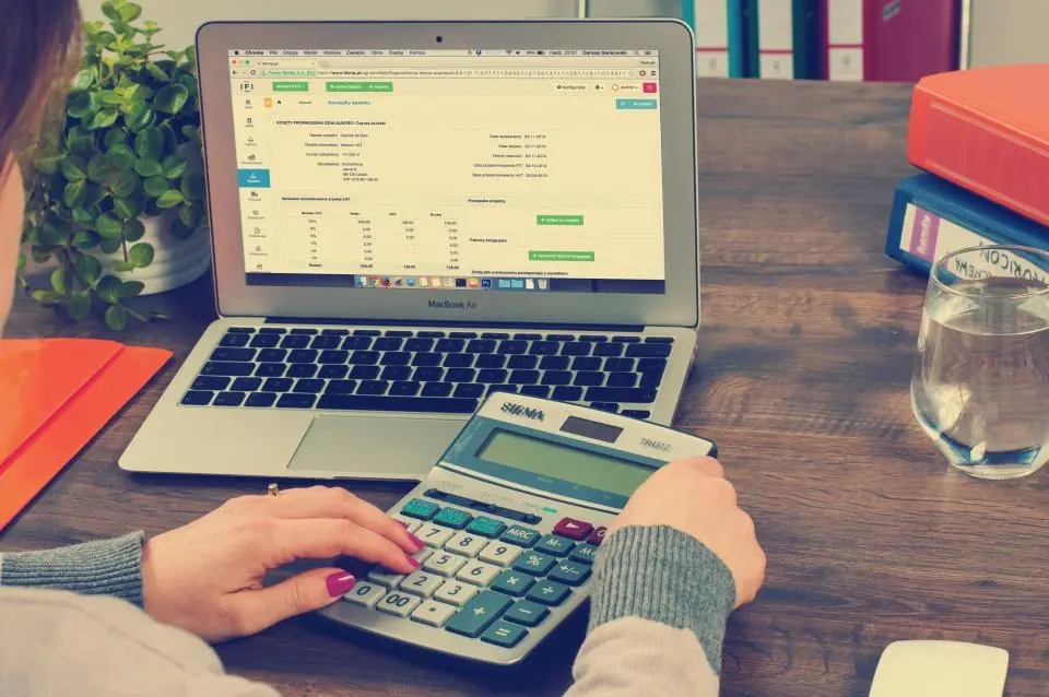 Mãos digitando em uma calculadora ao lado de um notebook com planilha aberta